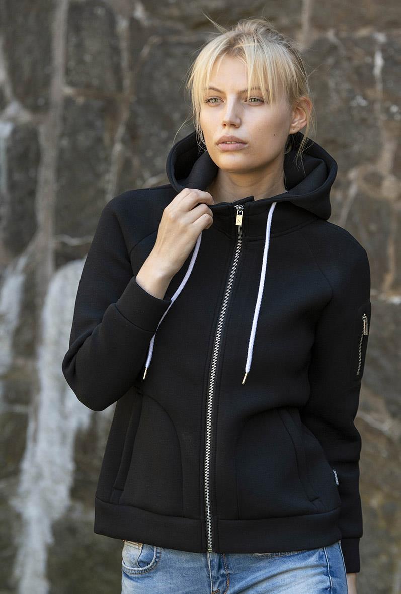 Kvinna i svart hoodie med dragkedja och vita snören, stående utomhus framför en stenvägg