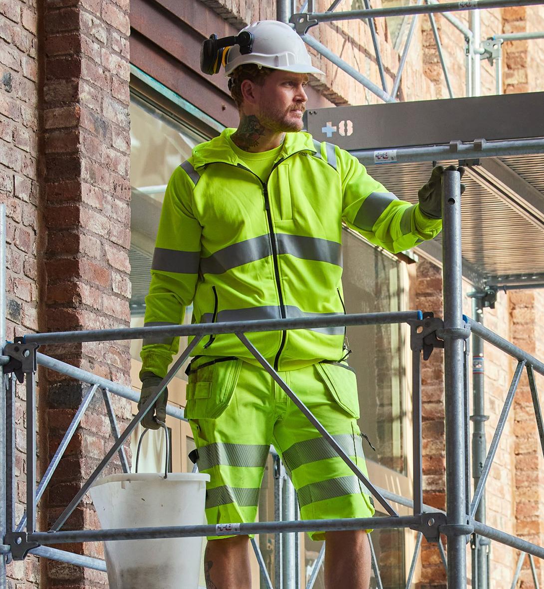 Man i högsynkig grön sweatjacka och shorts med reflexremsor på byggarbetsplats, står på scaffolding och tittar åt sidan
