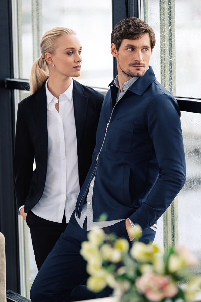 Två personer i formella kläder, en man i en svart overshirt och en kvinna i en vit skjorta och svart kavaj, står vid ett fönster.