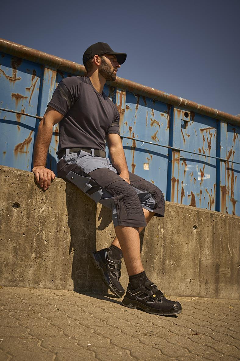 Man sitter på en betongmur med svart keps och sportskor, bakgrund av en blå, rostig metallbarriär