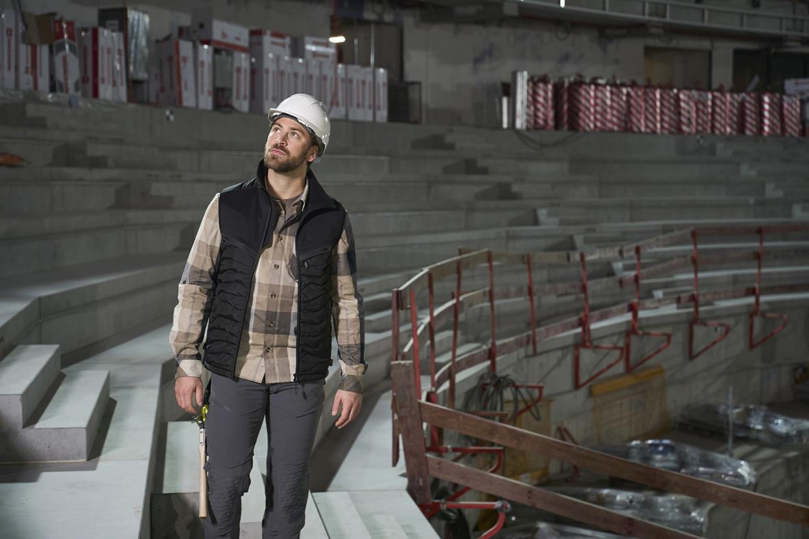 Man i mörk sand-rutig flanellskjorta och svart väst står i ett industriområde med byggnadsställningar och trappor