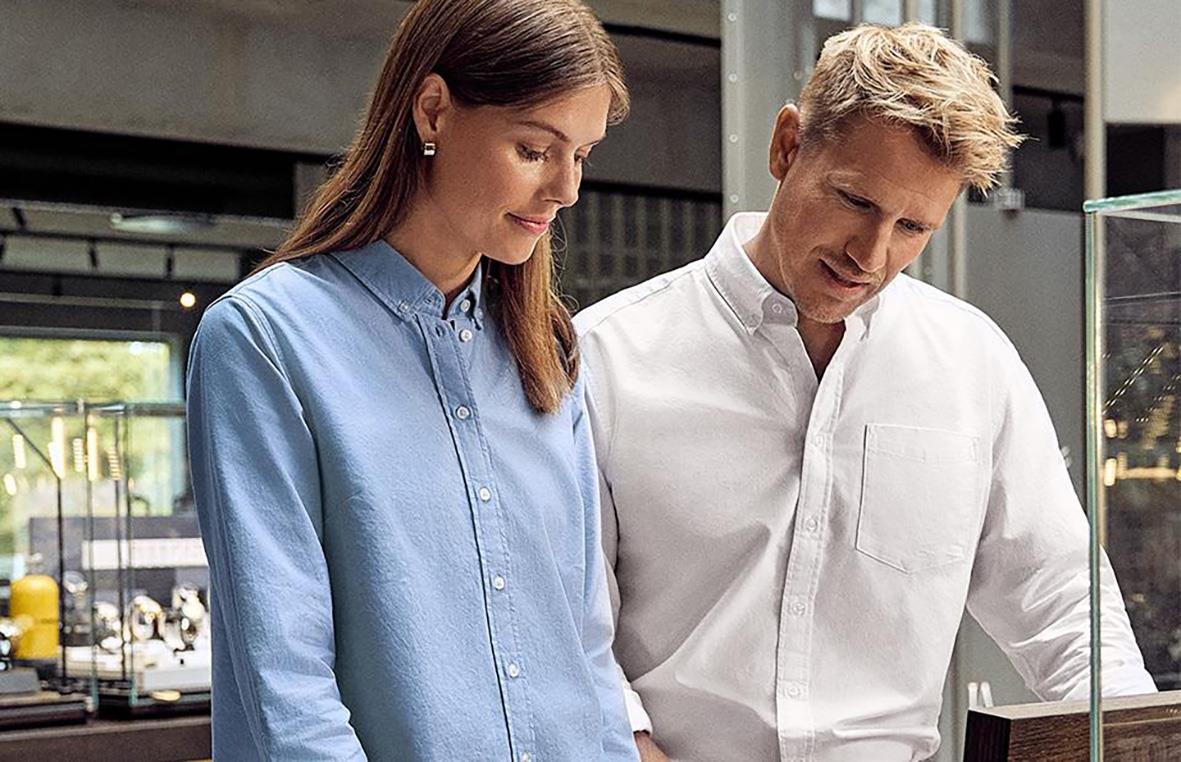 Två personer tittar på något, en kvinna i blå skjorta och en man i vit skjorta, i en modern inomhusmiljö