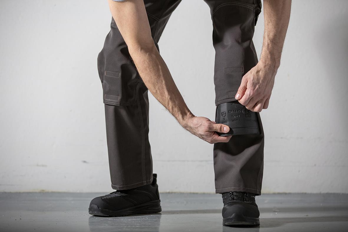 Person som justerar ett grått knäskydd på sitt vänstra knä, stående mot en ljus bakgrund