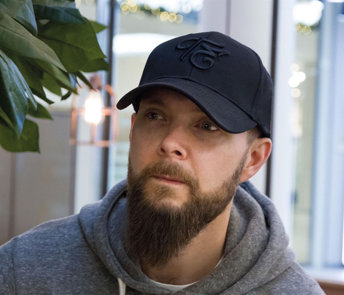 Man bär svart keps Trend Eagle 272 Stretch Svart, grå hoodie, inomhus med växter och fönster i bakgrunden