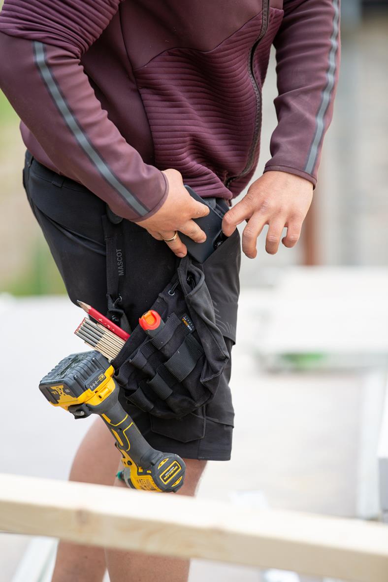 Hantverkare med svart verktygsväska och verktyg i bälte, klädd i vinröd jacka och svarta shorts