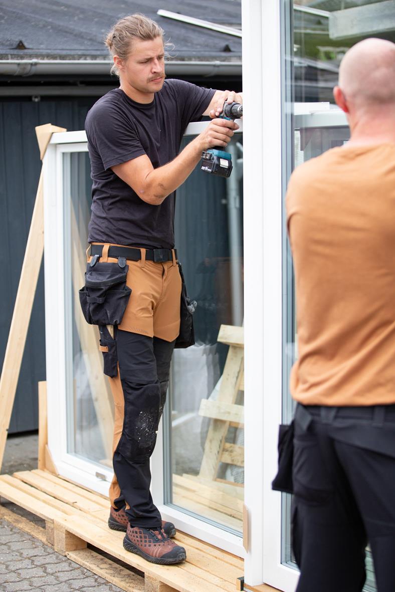 Man med svart t-shirt och bruna arbetsbyxor med verktygspåse, står bredvid ett fönster och använder en skruvdragare utomhus