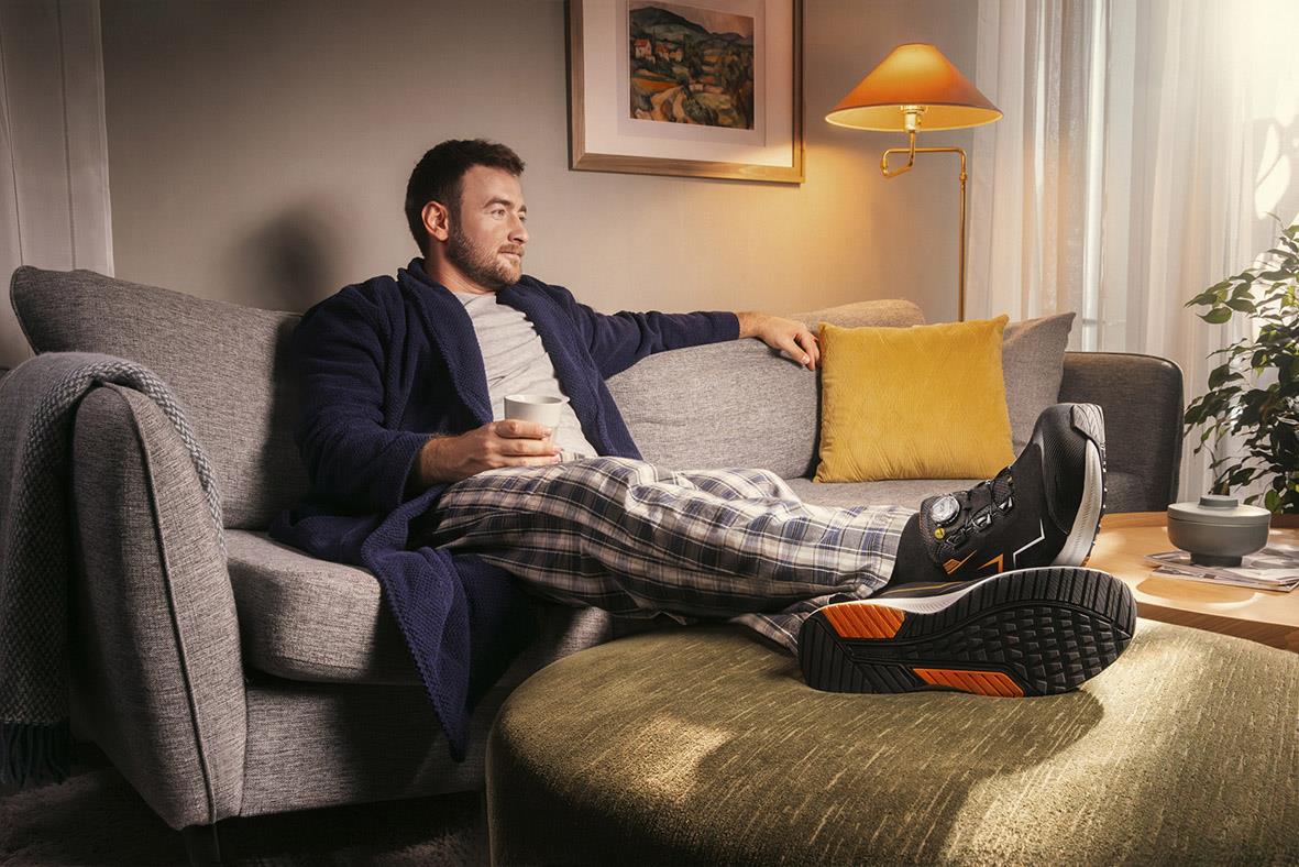 Man sitter på soffan med fötterna på en fotpall, bär skyddssko Monitor Recharge One 39, i ett vardagsrum