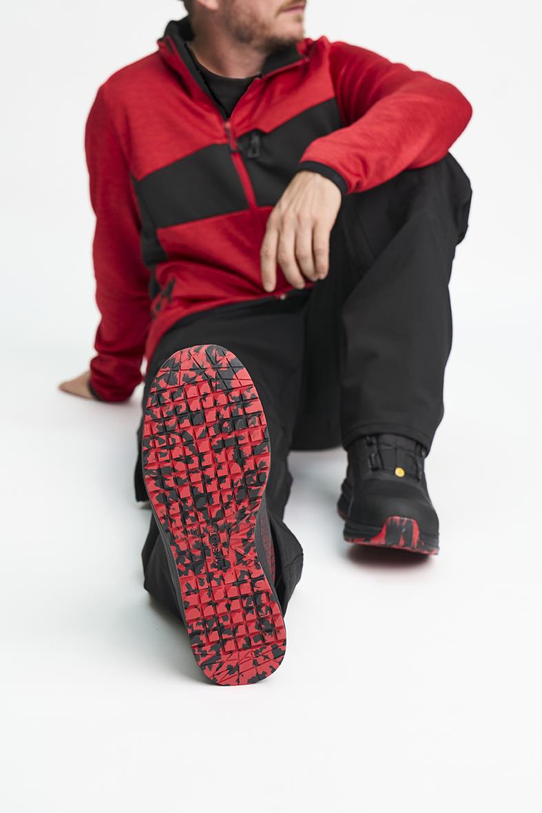 Person i röd och svart jacka visar svart skyddskänga med röd sula, sittande på golvet