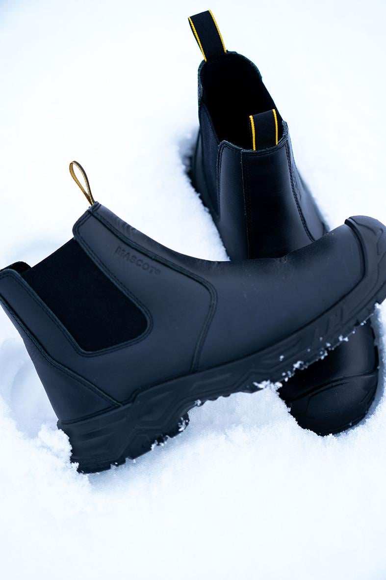 Svarta skyddskängor i gummi med tjock sula och dragöglor, placerade i snö