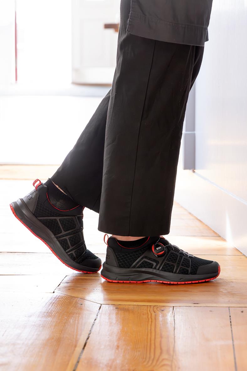 Person som bär svarta arbets- eller fritidsskor med röda detaljer, stående på trägolv