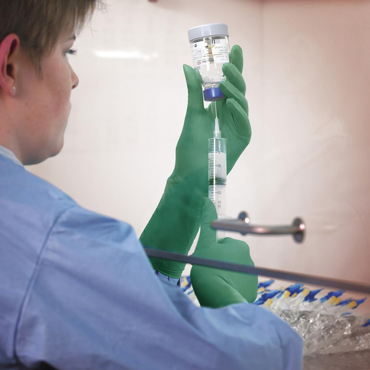 Person i blå arbetskläder och gröna handskar hanterar en spruta och ett provrör i ett laboratorium