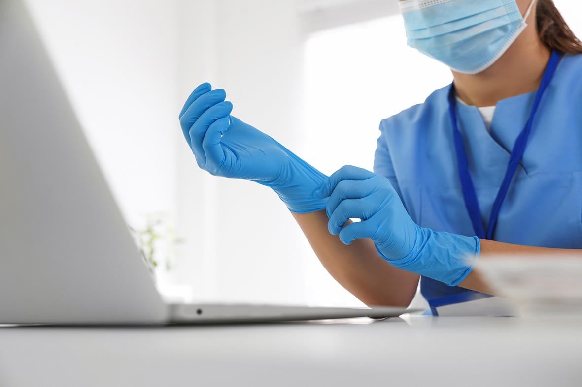 Person i blå skyddshandskar och munskydd som förbereder sig för arbete vid dator