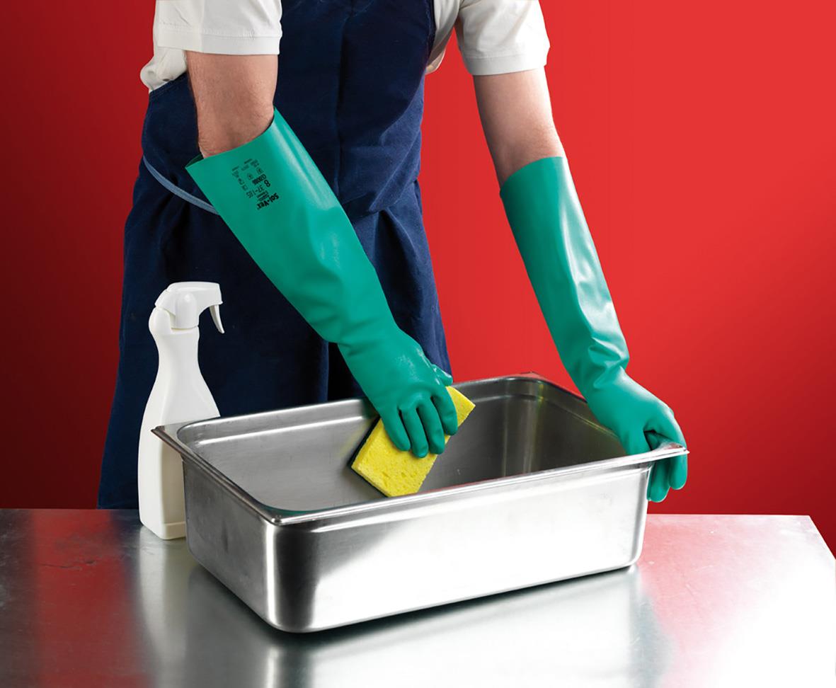 Person som rengör en metallbox med gröna kemskyddshandskar, sprayflaska och svamp mot en röd bakgrund