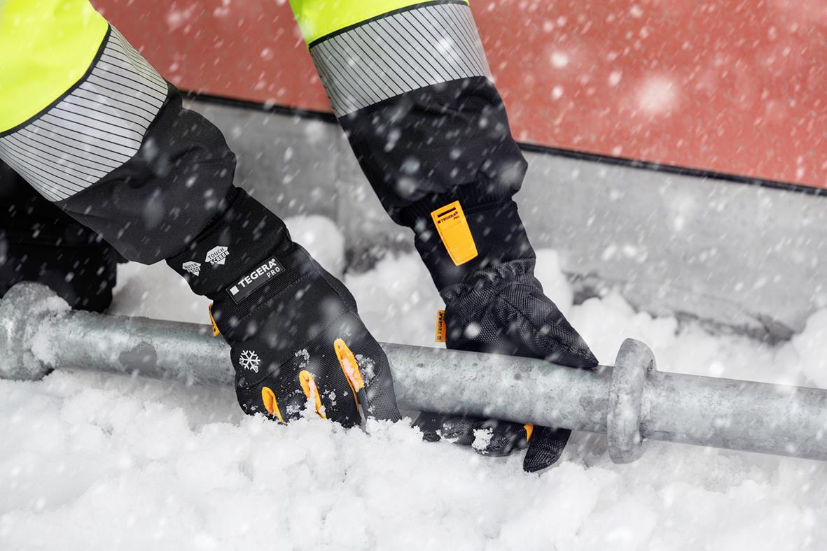 Vinterhandskar Tegera 9130 Svart används i snöväder, visar händer i svarta handskar med orange detaljer, som håller i ett metallrör.