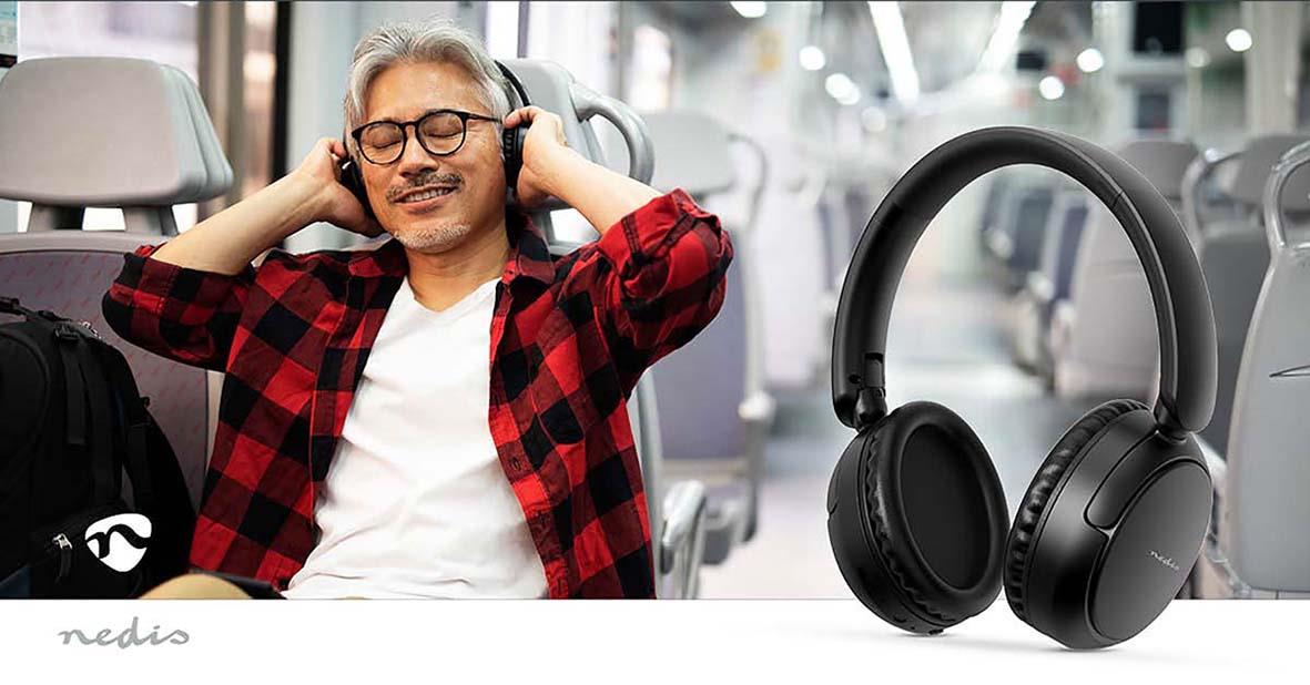 Man med grått hår och glas som njuter av att lyssna på musik med trådlösa svarta over-ear hörlurar i ett flygplan