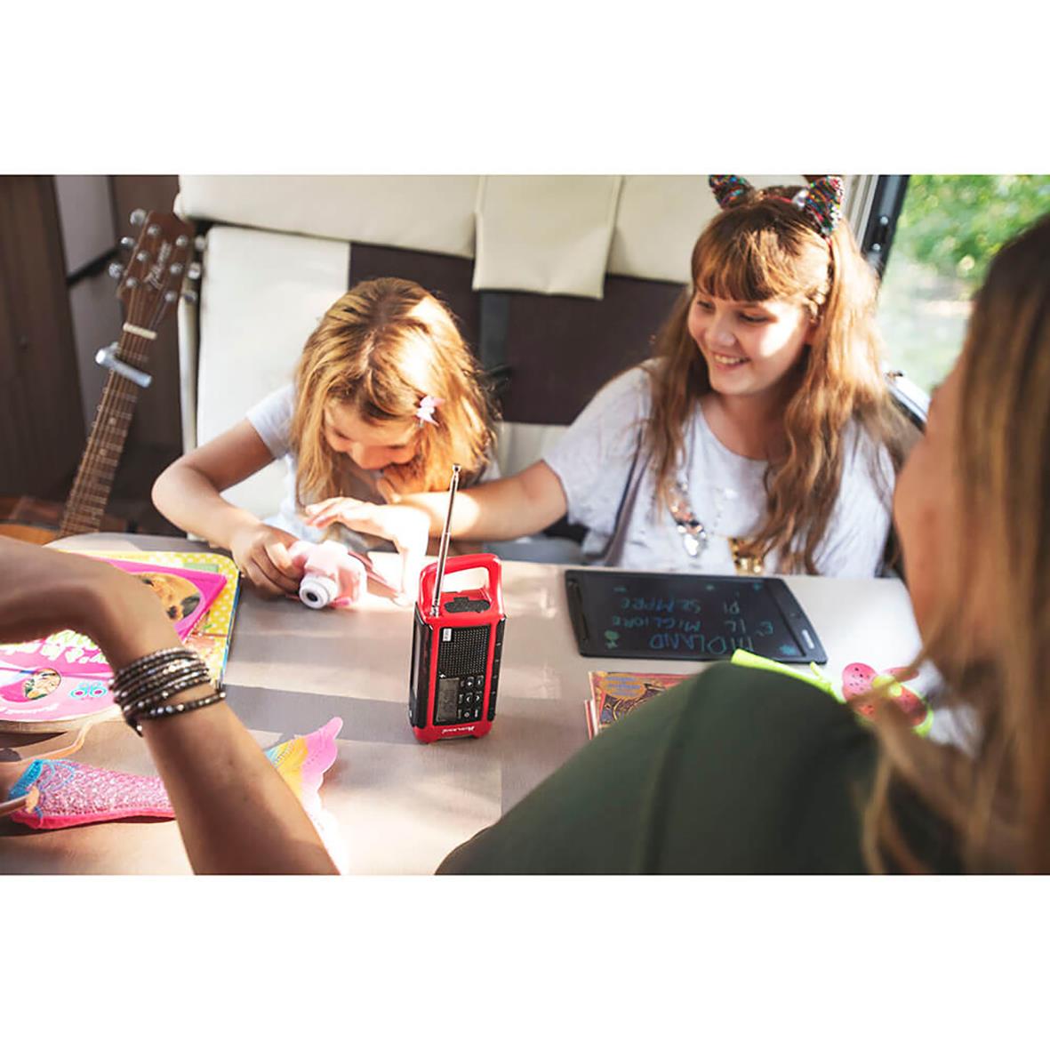 Två unga tjejer leker med en Midland nödradio med ficklampa och powerbank på ett bord, leende och glada i ett inomhusrum