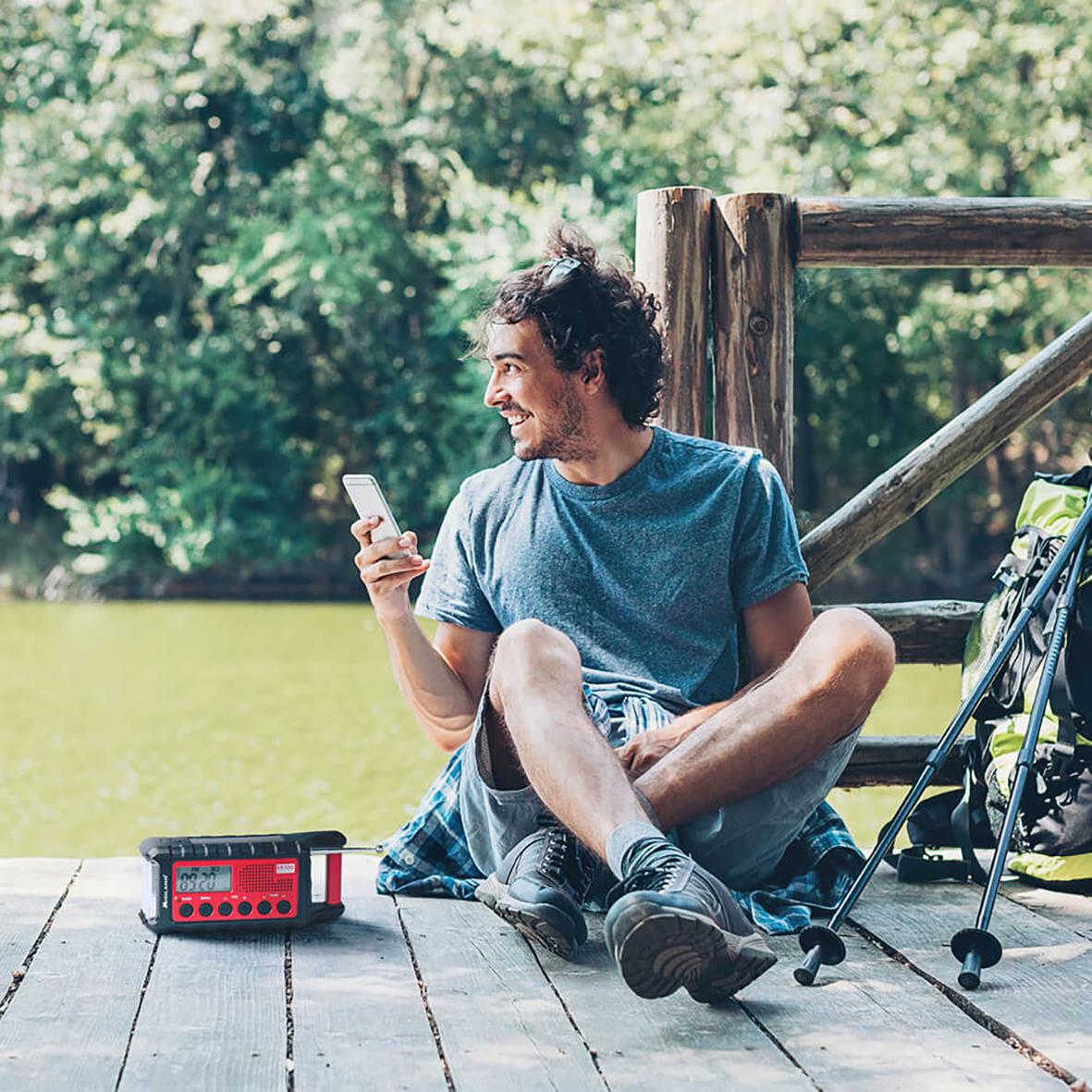 Man sitter på en brygga med en ficklampa och powerbank, omgiven av natur och en sjö, använder sin mobil