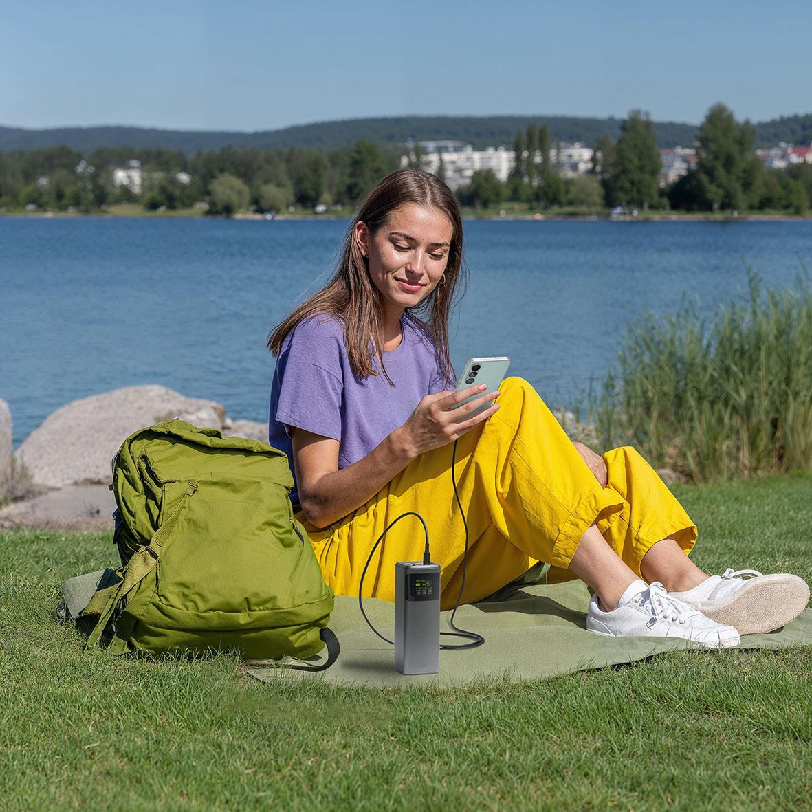 Kvinna sitter på gräset vid vattnet med mobiltelefon, ryggsäck och högtalare, njuter av en dag utomhus
