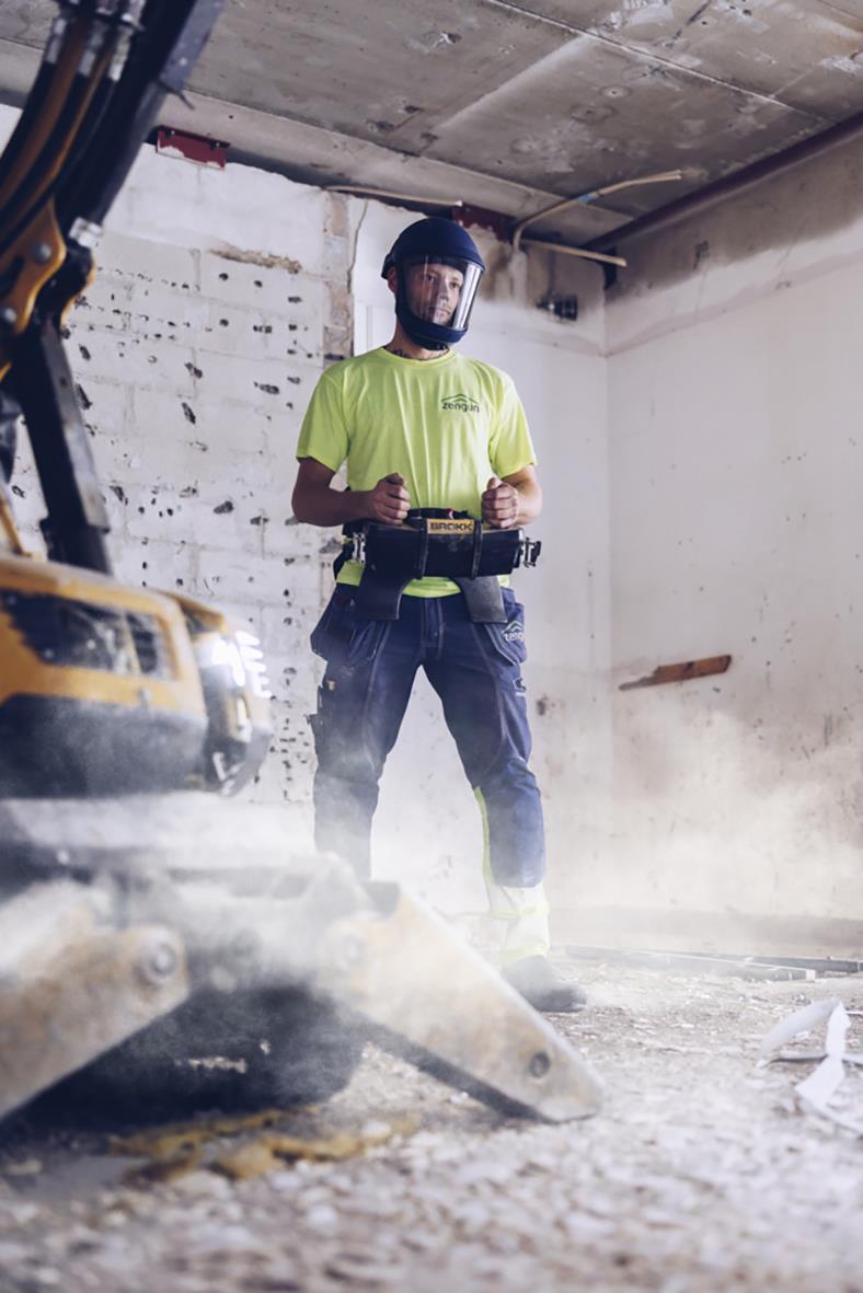 Person i byggarbetsmiljö med skyddshjälm och reflexväst, omgiven av byggmaterial och maskiner